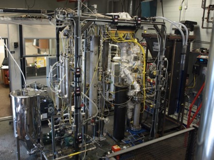 Installation industrielle complexe montrant l’usine pilote de pyrolyse rapide à lit fluidifié de CanmetÉNERGIE-Ottawa. L’image montre des tuyaux métalliques raccordés, des colonnes cylindriques et des composants isolés ainsi qu’une série de fils et de jauges de contrôle. À gauche, un grand réservoir en acier inoxydable est raccordé au système, tandis que plusieurs boyaux et capteurs sont reliés à l’unité centrale. 