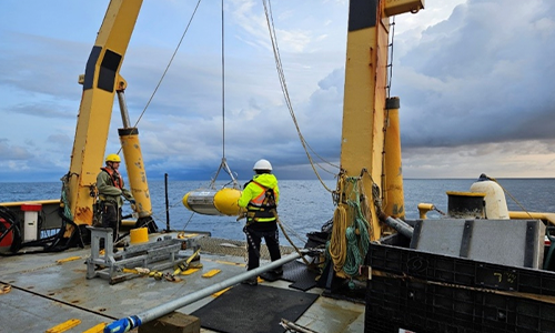 eux personnes portant des gilets de sécurité et des casques travaillent sur une plateforme extracôtière, entourée par la mer. Un grand objet est suspendu à des câbles près du bord de la plateforme. 