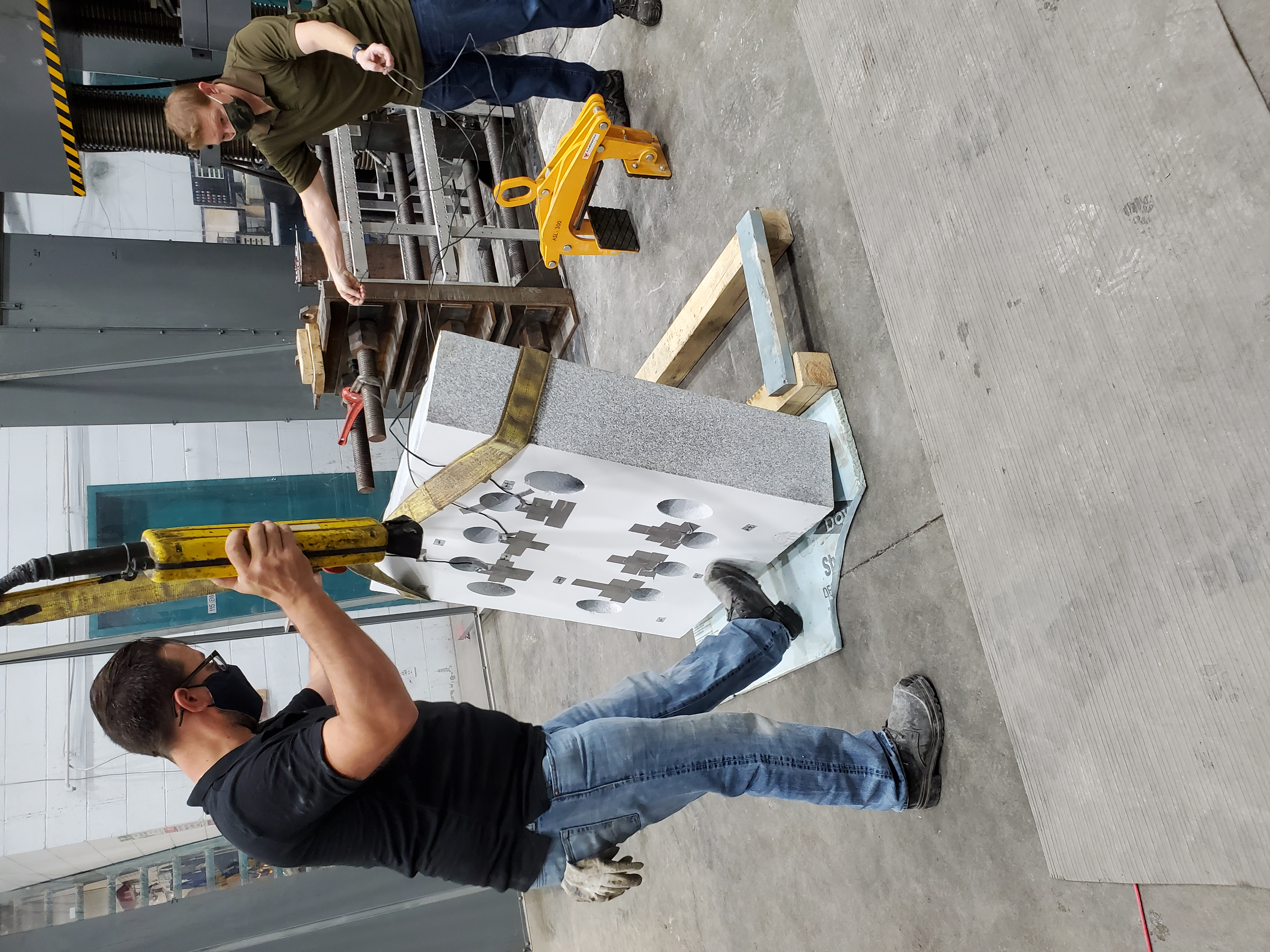 Une grande machine d’essai industrielle avec des colonnes hydrauliques verticales est placée derrière une barrière de protection transparente montée sur roulettes.