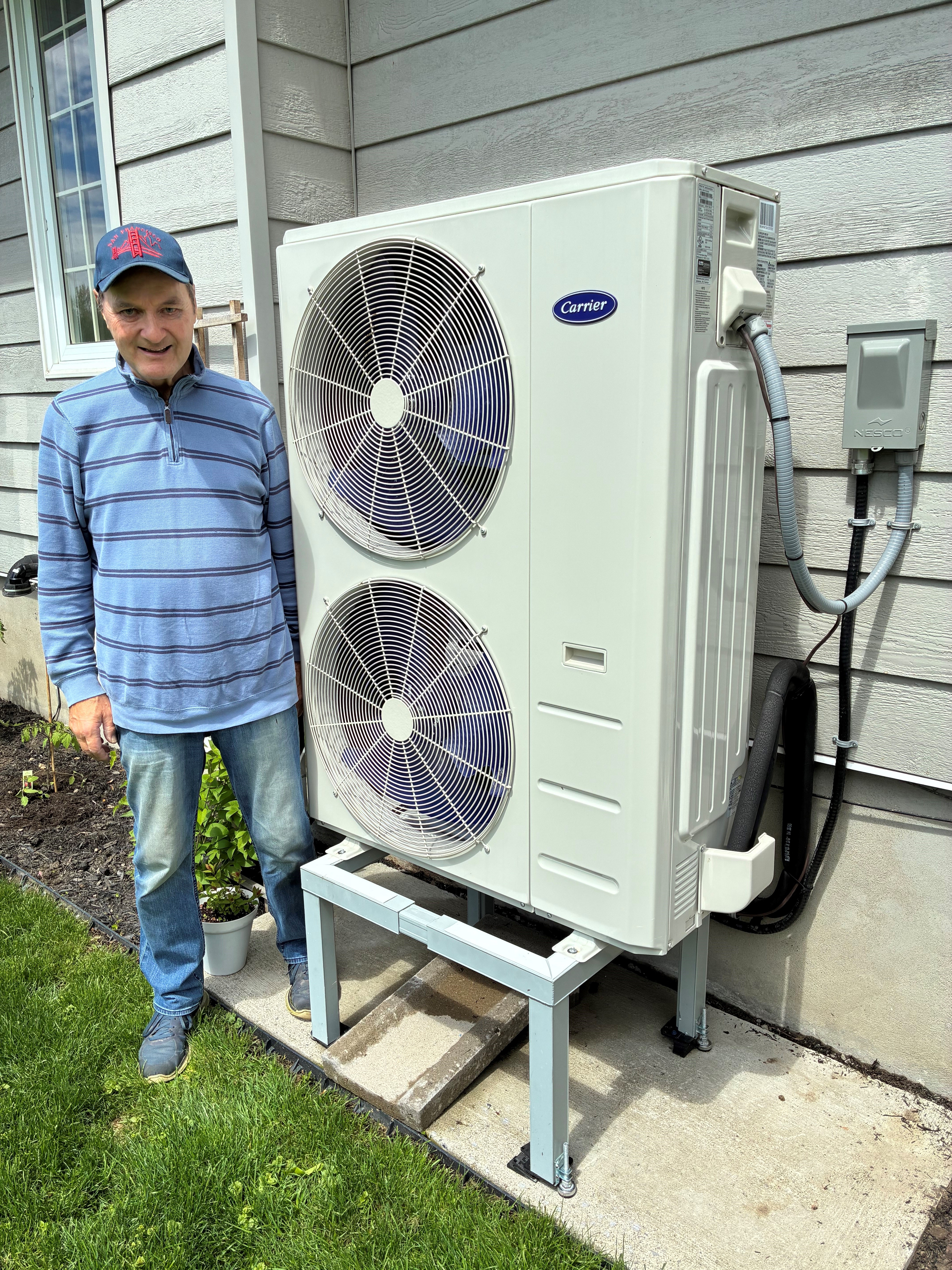 A smiling man neara heat pump on a metal stand next to his home