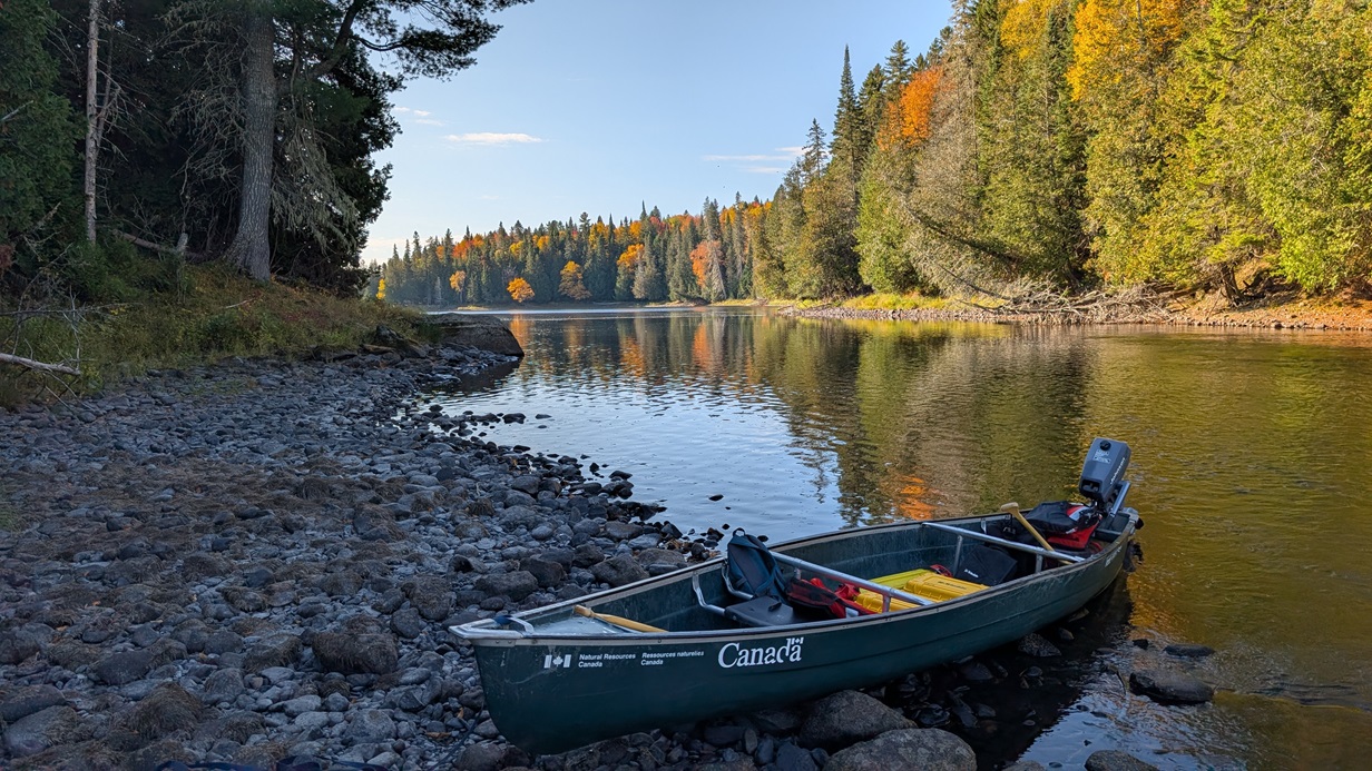 Un petit canoë vert du gouvernement du Canada est tiré sur la rive rocheuse d’une rivière, chargé de matériel de travail sur le terrain dans un environnement isolé.