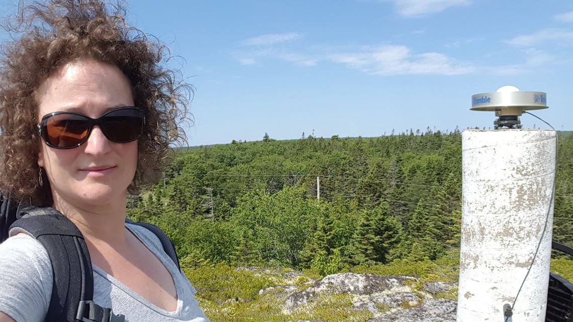 Une femme portant des lunettes de soleil se tient debout dans une forêt, près d’un cylindre en béton relié à une boîte noire contenant un récepteur de signaux satellitaires.