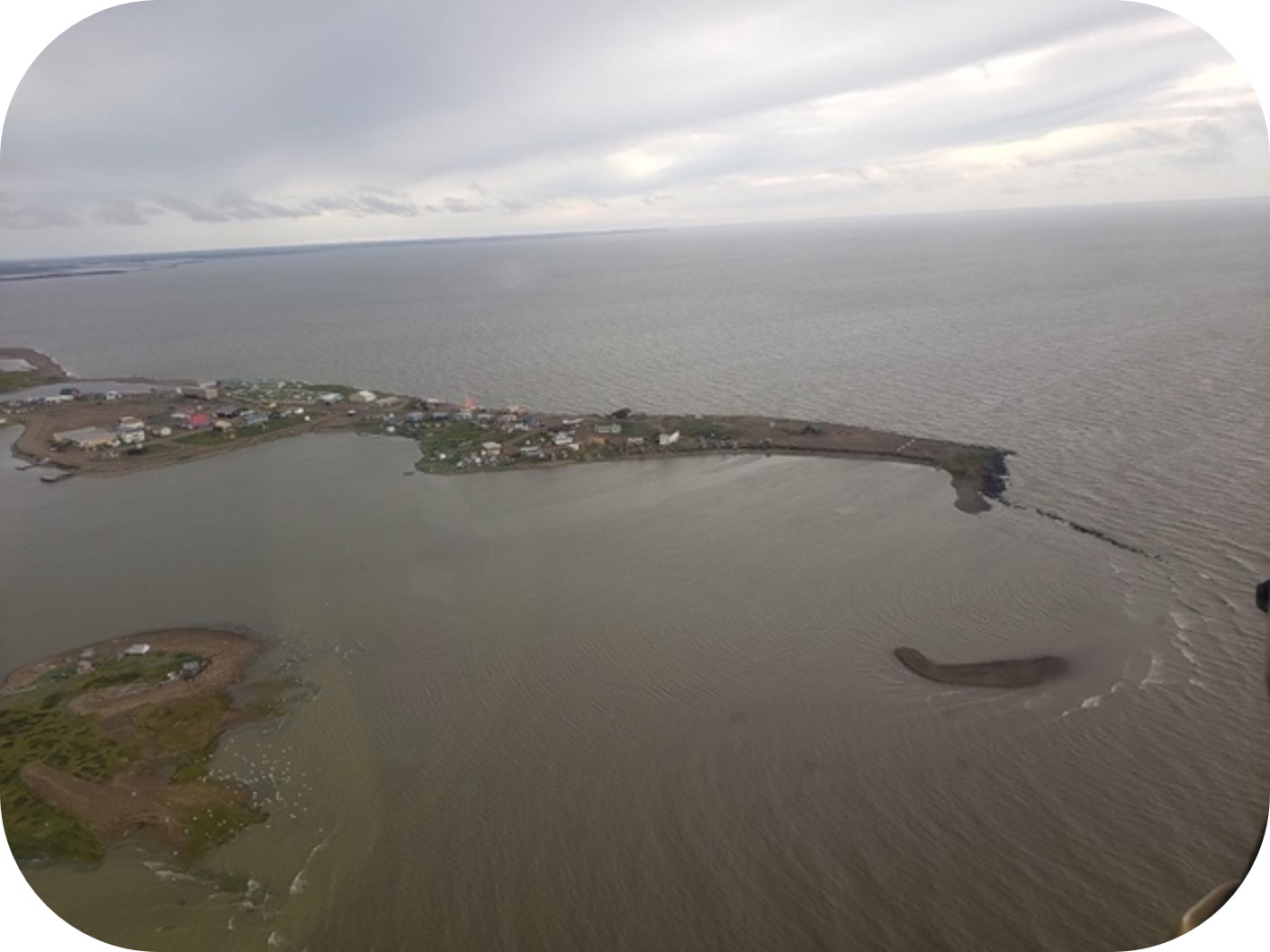 Vue aérienne d’une petite communauté côtière sur une péninsule étroite entourée d’eau sous un ciel couvert.