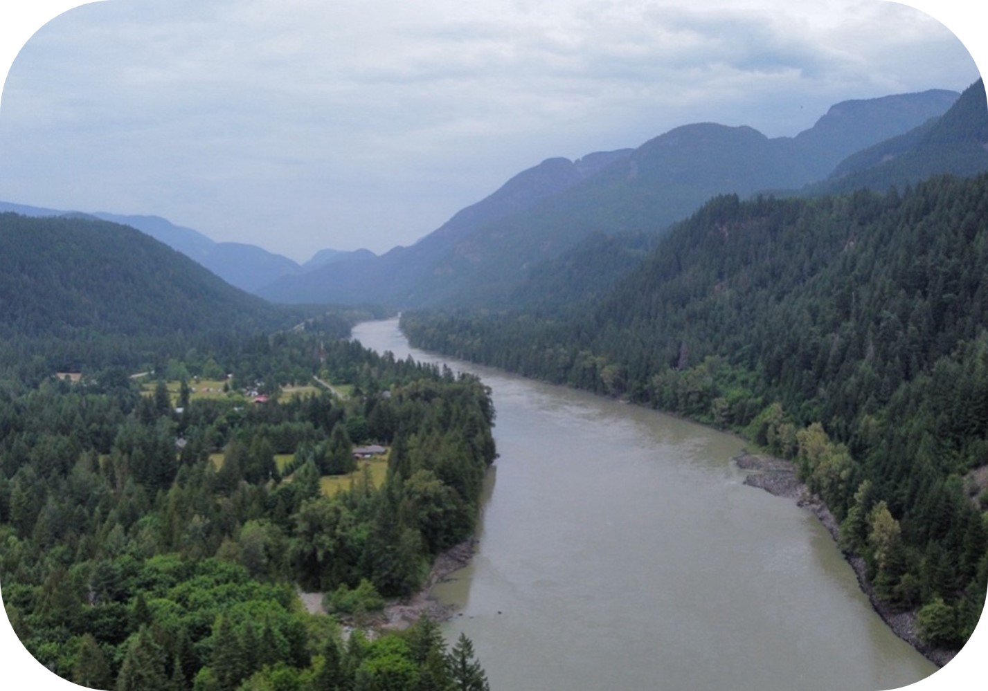 Vue aérienne d’un fleuve traversant une vallée montagneuse boisée avec des bâtiments ruraux sous un ciel nuageux.