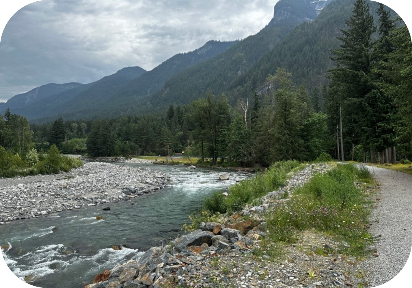 Rivière traversant une vallée montagneuse boisée, longée par un sentier de gravier sous un ciel couvert.