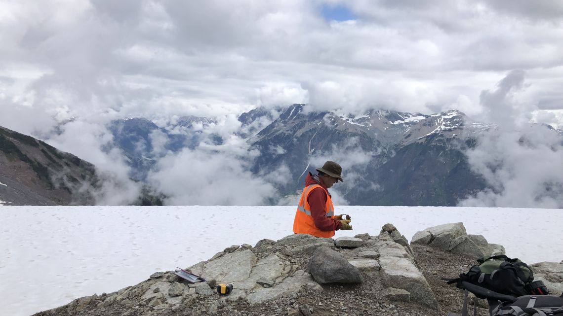 Un chercheur portant un gilet de sécurité orange examine un échantillon de roche sur un contrefort montagneux rocheux enneigé, devant des montagnes couvertes de nuages.