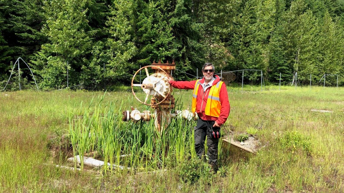 Steve Grasby, portant un gilet de sécurité orange, se tient debout à côté d’un ancien puits géothermique, dans une clairière herbeuse entourée de forêt.